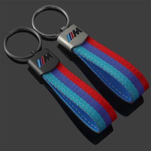 Pode incluir: Dois chaveiros com um anel de metal preto e uma alça com listras vermelhas, azuis e azul claro. O chaveiro tem uma placa de metal com as letras "IIM" em uma combinação de cores correspondente.