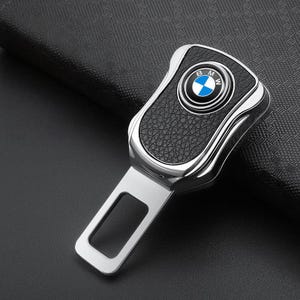 May include: A silver and black car seat belt buckle with the BMW logo. The buckle has a textured black center and a silver frame. The BMW logo is a blue and white circle with the letters BMW.