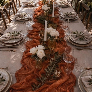 May include: An elegantly arranged table setting featuring a rust-coloured runner, white roses, and slender white candles. The table is set with plates, cutlery, and wine glasses, creating a sophisticated atmosphere for a special occasion or wedding.