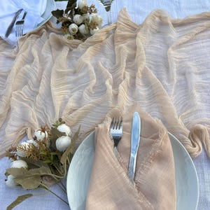 May include: A table setting featuring a beige gauze table runner, white plates, folded napkins, cutlery, and floral decorations. A martini glass is also visible. The scene is set outdoors.