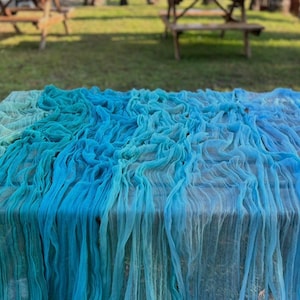 Op de afbeelding: Een rechthoekige tafel bedekt met een vloeiende, gelaagde stof in tinten blauw en teal. De stof drapeert over de tafel en creëert een textuur, oceaanachtige uitstraling. De tafel staat buiten, met banken en gras op de achtergrond.