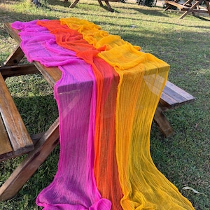 Peut inclure: Une table de pique-nique en bois ornée d'un tissu coloré et fluide. Le tissu passe du rose vif à l'orange puis au jaune. La matière texturée retombe sur les côtés de la table, créant une présentation attrayante.