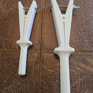 Może przedstawiać: Dwa białe plastikowe narzędzia do motków przędzy są pokazane na brązowym skórzanym tle. Narzędzia mają górną część w kształcie litery V z metalową śrubą i uchwytem. Jeden jest mniejszy od drugiego.