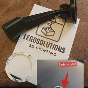 May include: Black 3D-printed car phone mount with a circular base and adjustable arm. Includes a white instruction sheet with the text "LEGOSOLUTIONS 3D PRINTING". Also shown is a sticker and a close-up of a button with the text "Slots in here".