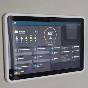 May include: A wall-mounted smart home control tablet with a white frame and a black screen displaying a dark-themed dashboard. The interface shows weather information, a thermostat set to 65 degrees Fahrenheit, and various home automation controls.