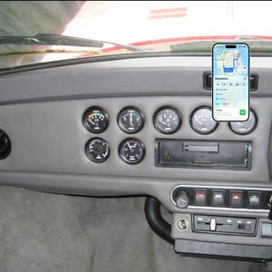 May include: A grey car dashboard featuring a series of circular analogue gauges, a central radio slot, and a smartphone mounted on the right side displaying a navigation map with directions and a 27-minute estimated arrival time on the screen.