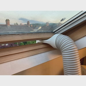 Puede incluir: Un tubo de escape flexible y ondulado de plástico blanco para aire acondicionado portátil, con una boquilla plana y ancha que está colocada de forma segura dentro de la abertura de un marco de ventana de techo de madera.
