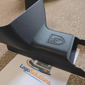 Puede incluir: Un componente de plástico negro con superficie texturizada y un logotipo. El componente tiene una forma curva con una base rectangular y dos extensiones puntiagudas. El logotipo es un escudo con un diseño detallado. El componente descansa sobre un papel con el texto "Lego Solutions".