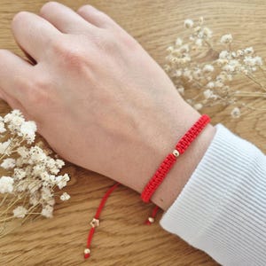 Könnte beinhalten: Ein rotes Armband aus geflochtener Kordel mit einer einzelnen kleinen goldfarbenen Perle in der Mitte. Das Armband hat zwei herabhängende Enden, die jeweils mit einer winzigen goldfarbenen Perle versehen sind, eine davon sternförmig.