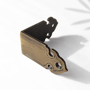 May include: A bronze-colored metal corner bracket with decorative cutouts and a right-angle shape. The bracket has a vintage aesthetic, suitable for furniture or decorative applications. The metal has a slightly aged appearance.