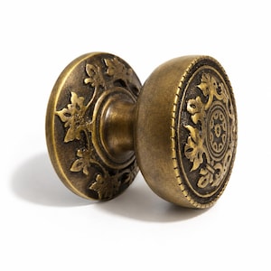 May include: A round brass door knob with an antique finish, featuring a raised relief pattern of stylized leaves and a central circular motif. The knob has a textured surface with a beaded border around the edge and a cylindrical central stem.