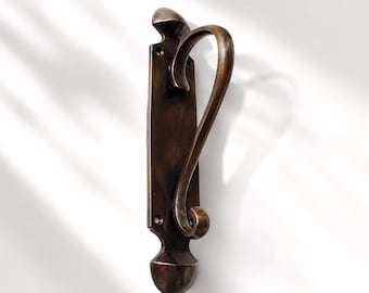 May include: A dark bronze-toned metal door handle featuring a vertical rectangular backplate with rounded ends and two screw holes. The handle has a curved, sweeping design that extends from the top and loops back to the center of the plate.