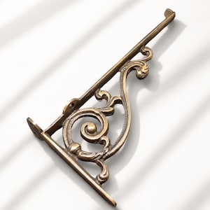 May include: A bronze-toned metal shelf bracket featuring an L-shaped frame with a decorative scrollwork design. The bracket includes two mounting points with pre-drilled holes at the base and a small hook at the top end of the diagonal support arm.