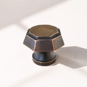 May include: A bronze-toned cabinet knob with an octagonal top and a cylindrical base. The knob has a geometric design and a textured surface, suitable for furniture hardware. The finish has a vintage or antique appearance.