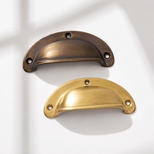May include: Two semi-circular cup drawer pulls with a curved grip. One pull features a dark, aged bronze finish, while the other has a bright, brushed brass finish. Each handle includes three pre-drilled screw holes for mounting purposes.