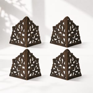 May include: Four decorative corner brackets with an ornate, openwork design. The brackets are made of a dark brown metal and feature a repeating floral and scroll pattern. Each bracket has a right-angle shape with pre-drilled holes for mounting.