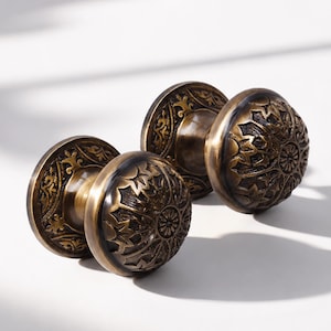 Peut inclure: Deux poignées de porte rondes avec une finition en laiton antique foncé. Chaque poignée présente un motif floral et végétal en relief détaillé sur la face circulaire et la plaque arrière assortie, créant un aspect métallique texturé.
