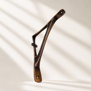 May include: A triangular metal shelf bracket with a dark bronze finish. The bracket features a slender, angled support arm with a decorative cutout design and two circular mounting holes at the top and bottom ends for secure wall attachment.
