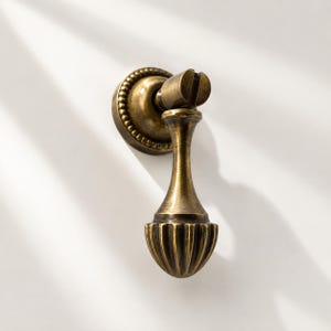 May include: A brass-toned metal drop pull handle featuring a circular backplate with a beaded edge and a vertical, teardrop-shaped pendant with a fluted, ribbed base. The hardware has a weathered, antique finish with dark highlights.