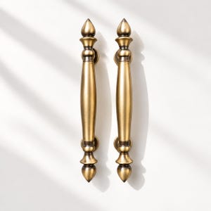 May include: Two vertical door handles with a brushed brass finish. Each handle features a central cylindrical shaft that tapers toward the ends, topped and bottomed with decorative finials and circular mounting plates for secure attachment.