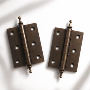 Peut inclure: Deux charnières de porte rectangulaires en métal de couleur bronze, dotées d'un cylindre central à cinq articulations et d'embouts décoratifs en haut et en bas. Chaque plaque de charnière comporte trois trous de vis circulaires alignés.