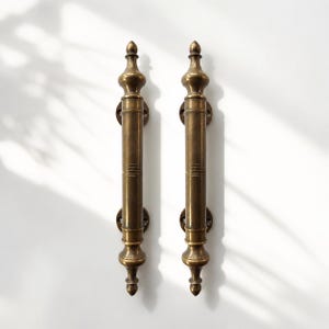 May include: A pair of vertical brass door handles featuring a cylindrical central shaft with horizontal etched lines, topped and bottomed by decorative finials, and secured to the surface by two circular mounting plates on each handle.