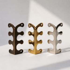 May include: Three decorative metal corner brackets in bronze, gold, and silver. Each bracket has a unique, stylized design with rounded edges and three holes for mounting. The brackets are displayed on a white surface.