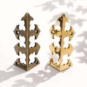 May include: Two decorative metal corner brackets. One is a bronze color, and the other is gold. Both have an ornate, openwork design with a triangular shape, perfect for furniture or craft projects.