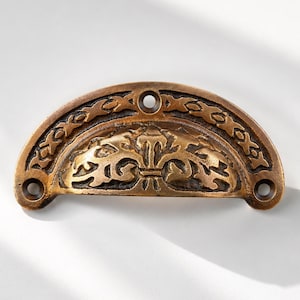 May include: A semi-circular bronze-toned metal drawer pull featuring an embossed floral pattern in the center and a repeating geometric cross design along the curved top edge. The hardware includes three circular screw holes for mounting purposes.