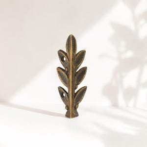 May include: A bronze-colored metal leaf with a textured surface. The leaf has a series of pointed, oval-shaped leaves along a central stem. The stem has three small holes. The leaf is standing upright on a white surface.