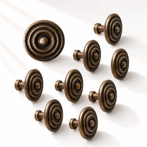 Könnte beinhalten: Eine Sammlung von neun runden Schrankknöpfen mit einem konzentrischen Kreismuster. Jeder Knopf ist in einem gealterten Bronzeton mit einer dunklen, verwitterten Patina gehalten und weist einen erhöhten Mittelpunkt sowie einen kurzen Schaft auf.