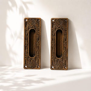 Peut inclure: Deux poignées de porte encastrées rectangulaires en métal couleur bronze, ornées d'un motif en relief de volutes entourant une prise ovale centrale. Chaque poignée comporte quatre trous de vis circulaires, un à chaque coin, pour la fixation.