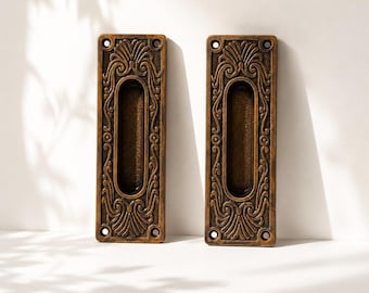May include: Two rectangular bronze-toned metal flush door pulls featuring an ornate, raised scrollwork pattern surrounding a central oval recessed grip. Each pull has four circular screw holes, one at each corner, for mounting onto a door surface.