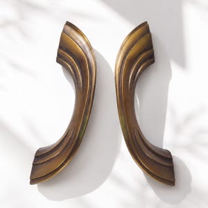May include: Two curved door handles made of brushed bronze metal. Each handle features a series of layered, stepped ridges along its length, creating a smooth, sweeping arc shape with a dark, antiqued finish and a metallic sheen.