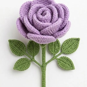 Puede incluir: Una rosa de ganchillo hecha a mano en un suave tono lavanda, con pétalos superpuestos y tejidos apretados. La flor está unida a un tallo verde resistente con cuatro hojas verdes texturizadas distintas a los lados, todo hecho de hilo.