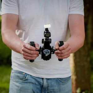 May include: A black camera stabilizer rig featuring two side handles, a central mount for a small action camera, and a small rectangular LED light attached to the top. The device includes a fuzzy microphone windscreen mounted on the front center.