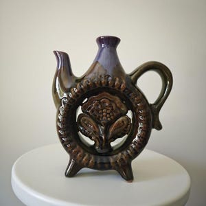 Könnte beinhalten: Dekorative Keramikkrug mit dunkelbrauner und violetter Glasur. Der Krug hat ein kreisförmiges Design mit einem floralen Motiv in der Mitte, einen Griff, einen Ausguss und drei kleine Füße. Der Krug steht auf einem weißen Sockel.