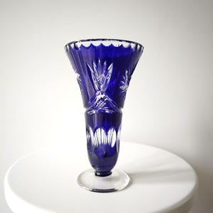 Könnte beinhalten: Eine kobaltblaue Kristallvase mit ausgestelltem Oberteil und klarer, kreisförmiger Basis. Die Vase weist aufwendige Schliffmuster und einen gewellten Rand auf. Die Vase ist etwa 25 cm hoch.