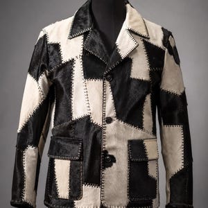 May include: A jacket constructed from a patchwork of black and white hair-on hide panels. The garment features a notched lapel, a three-button front closure, and two lower flap pockets. Each panel is joined by visible, thick, dark-colored stitching.