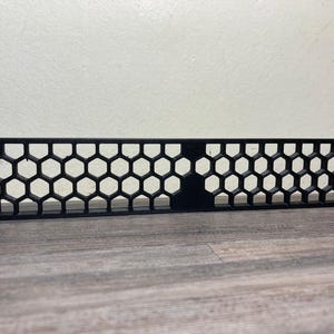 May include: Black metal rack panel with a honeycomb pattern. The panel has mounting holes on each end. The design features a series of hexagonal cutouts, creating an open, airy appearance. The panel is designed for equipment mounting.