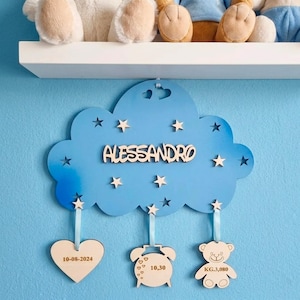 May include: A blue cloud-shaped wooden wall decoration with the name "ALESSANDRO" in wooden letters. It has star cutouts and three hanging wooden ornaments: a heart with "10-08-2024", a clock with "10,30", and a teddy bear with "KG.3,080".