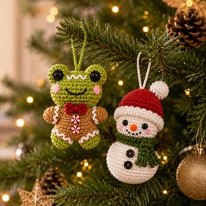 May include: Two crocheted ornaments hanging from a tree. One is a green frog with a brown gingerbread body, red bow tie, and white peppermint buttons. The other is a white snowman wearing a red hat with a white pom-pom and a green scarf.
