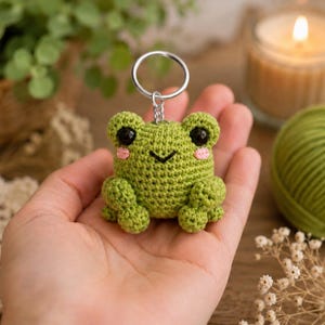 May include: A small, green, crocheted frog keychain featuring a round body, two prominent black eyes, a stitched black mouth, and two small pink circles on the cheeks. The frog is attached to a silver-colored metal split ring by a short chain.