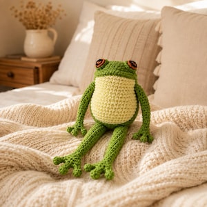 May include: A handmade crochet frog toy featuring a vibrant green body with a pale yellow belly and prominent orange and black eyes. The stuffed animal has long, slender arms and legs with detailed webbed feet, crafted from textured yarn.