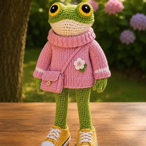 May include: A crocheted frog doll with green skin, large black and yellow eyes, and a cream-colored chin. The frog wears a pink knit turtleneck sweater with a white flower appliqué, a matching pink crossbody bag, and yellow sneakers with white toes.