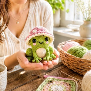 May include: A small, green, crocheted frog plush toy wearing a pink mushroom cap with white spots. The frog has black plastic eyes, a stitched smile, and pink blush marks on its cheeks. The body is made of yarn with a cream-coloured belly.