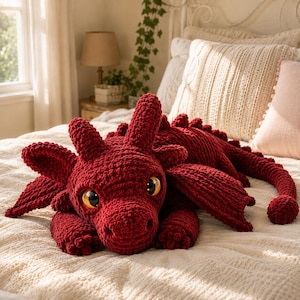 May include: A large, deep red crochet dragon plush toy featuring a textured yarn surface, two prominent horns on its head, wide wings, and large, glossy amber-colored eyes. The creature has a long, segmented tail and small clawed feet.