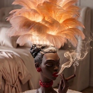 Può includere: Una lampada da tavolo raffigurante un busto dalla pelle scura con un'acconciatura a chignon alto, che indossa orecchini, un girocollo e un braccialetto a forma di rose rosse. Il paralume è fatto di piume rosa chiaro. La figura tiene un sigaro.