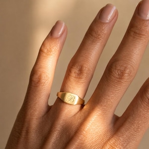May include: A gold signet ring with the letter "B" engraved on the rectangular face. The ring is worn on a finger, showcasing its polished, reflective surface. The ring's design is simple and elegant.