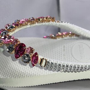 May include: A white flip-flop featuring a strap embellished with a variety of pink and dark gray faceted crystals in different shapes. The base of the strap includes a row of small, clear, round rhinestones set in a metallic frame.